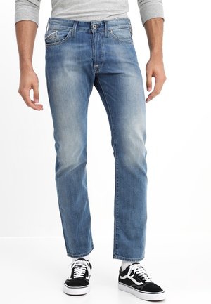 Man wearing faded blue jeans, gray long-sleeve shirt, white socks, and black-and-white sneakers standing against a white background.