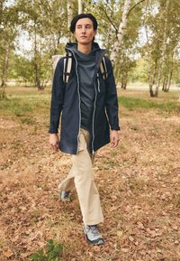 Navy waterproof jacket with a hood, gray patterned shirt, beige pants, and gray hiking shoes. Backpack with two shoulder straps.