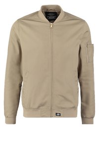 Veste bomber beige avec fermeture éclair avant, col et poignets côtelés, deux poches latérales et petite poche zippée sur la manche gauche.