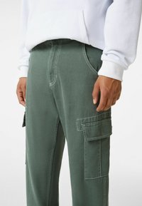 Gröna cargo-byxor i hållbar tyg, med sidofickor och kontrasterande vit sömnad, i kombination med en vit hoodie.