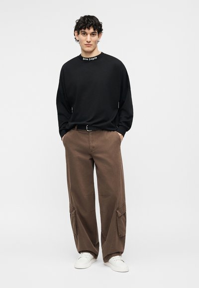 Uomo con capelli ricci che indossa una maglia nera a maniche lunghe con testo sul colletto, pantaloni cargo marroni, sneakers bianche, mani in tasca, in piedi dritto.