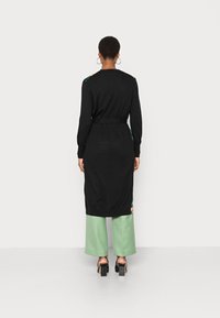 Zwarte gebreide jurk met lange mouwen, ceintuur op de taille en bloemaccenten op de schouders. Gecombineerd met mintgroene broek met wijde pijpen en zwarte hakken.