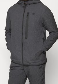 Man som bär en strukturerad mörkgrå Nike-hoodie och matchande byxor med synliga logotyper och en ficka med dragkedja på bröstet, med händerna i sidofickorna.