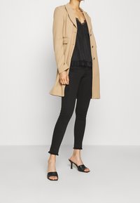 Abrigo largo beige, top negro a rayas con encaje en los bordes, jeans negros skinny con dobladillo deshilachado, mules negras de punta abierta, de pie contra un fondo blanco.