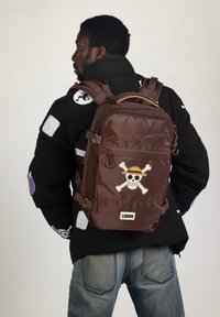 Mochila de nailon marrón con correas ajustables, gráfico de calavera y huesos cruzados en la parte frontal, dos bolsillos laterales y un asa en la parte superior.