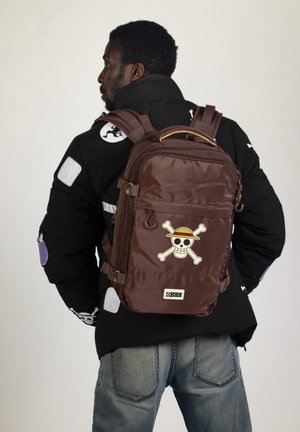 Mochila de nailon marrón con correas ajustables, gráfico de calavera y huesos cruzados en la parte frontal, dos bolsillos laterales y un asa en la parte superior.