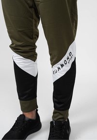 Olivově zelené sportovní kalhoty s černými a bílými geometrickými bočními panely. Mají elastické manžety a potisk loga na bílém panelu.