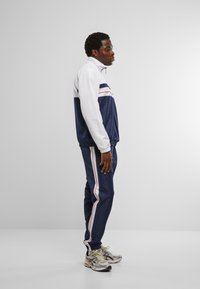 Marineblå og hvit joggebuksejakke med røde striper, kombinert med matchende bukser. Laget av glatt stoff, med elastiske mansjetter og sporty design.