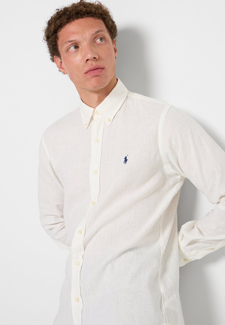 Chemise en lin de couleur crème avec un col boutonné, des manches longues et un petit logo bleu sur la poitrine. Tissu texturé et coupe classique.