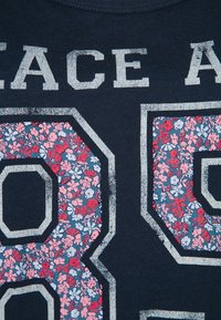 Námořnická modrá bavlněná trička s velkými květinovými číslicemi "8" a "0" v růžové a modré barvě, s textem "PEACE" v opotřebované bílé nad nimi.