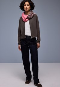 Dunkelgraues Sweatshirt mit Reißverschluss, weißes T-Shirt, dunkle Jeans mit kontrastierender Naht und ein rosa Schal mit Tiermuster. Braune Schuhe mit weißen Schnürsenkeln.