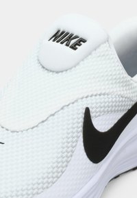 Baskets blanches sans lacets avec tige en mesh texturé, logo Nike noir et accent noir incurvé, dotées d'une semelle coussinée et d'un design épuré.