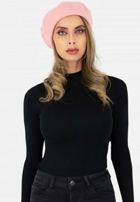Beret en laine rose sur de longs cheveux ondulés. Le modèle porte un shirt noir ajusté à manches longues et un jean sombre, avec un fond neutre.
