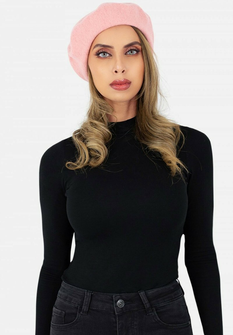Beret en laine rose sur de longs cheveux ondulés. Le modèle porte un shirt noir ajusté à manches longues et un jean sombre, avec un fond neutre.