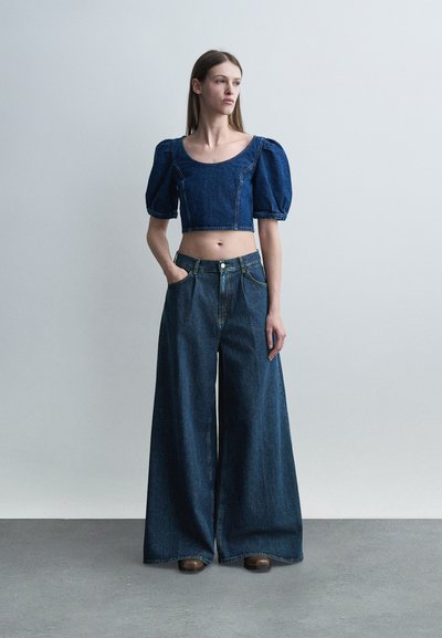 Γυναίκα står med το χέρι στην τσέπη, φορώντας cropped denim τοπ με φουσκωτά μανίκια και φαρδιά, ψηλόμεση denim παντελόνια, μπροστά σε απλό φόντο.