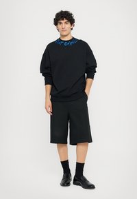 Sweatshirt preto com detalhes em texto azul na gola, combinado com calções pretos até ao joelho e sapatos pretos. Textura de tecido suave.
