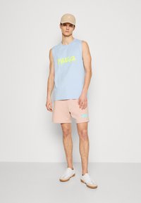 T-shirt sans manches bleu clair avec un graphique jaune, associé à un short rose avec un logo bleu. Baskets blanches avec des semelles marron clair.