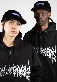 Sudaderas negras con cremallera y texto gráfico blanco, acompañadas de gorras de pana negras con logos azules, ambas con un diseño casual y moderno.