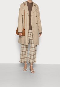 Trench-coat beige avec des boutons, pull en tricot marron, pantalon large à carreaux en crème et marron, et escarpins légers à lanières. Sac bandoulière marron.