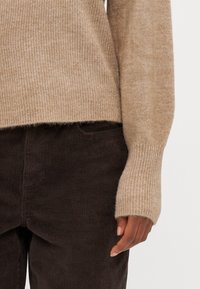 Manica di un maglione beige a coste con una consistenza morbida, abbinata a pantaloni in velluto a coste marrone scuro che mostrano un motivo sottile.
