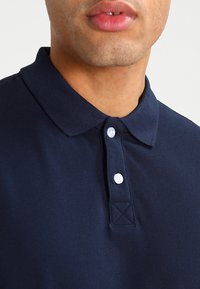 Marineblå polo skjorte med krage og tre hvite knapper. Har en teksturert stoff og trekantet søm detaljer ved kragen.
