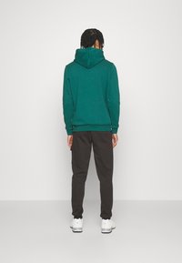Zelená mikina s kapucí na šňůrku, spojená s tmavými kalhotami. Nabízí volný střih a žebrované manžety. Bílé tenisky dotvářejí outfit.