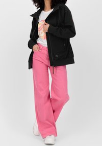 Persona che indossa una giacca nera con zip, una maglietta bianca con grafica, pantaloni larghi rosa e sneakers bianche, in piedi con una mano in tasca.