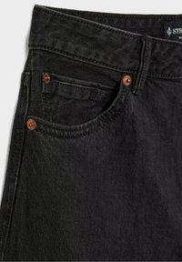 Gros plan sur la poche avant d'un jean en denim noir avec des rivets en cuivre et une boucle de ceinture visible à la taille.