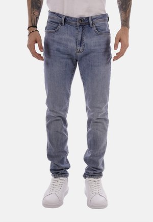 Uomo con jeans slim fit azzurri chiari, sneaker bianche e camicia bianca, in piedi con le braccia rilassate lungo i fianchi su sfondo bianco.