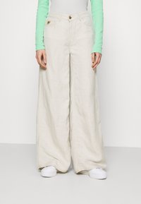 Béžové široké lněné kalhoty s vysokým pasem, které mají přední zapínání na knoflík a dvě přední kapsy, doplněné bílými teniskami.