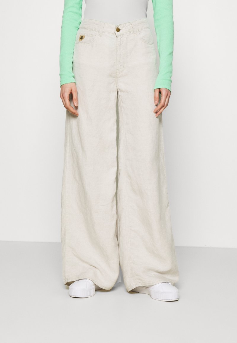 Béžové široké lněné kalhoty s vysokým pasem, které mají přední zapínání na knoflík a dvě přední kapsy, doplněné bílými teniskami.