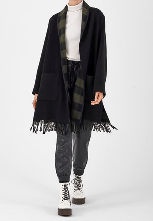 Kvinde iført sort frynset sjal med grønt ternet foer, hvid rullekrave, sorte bukser med snøre og hvide snørebåndsplatformstøvler.