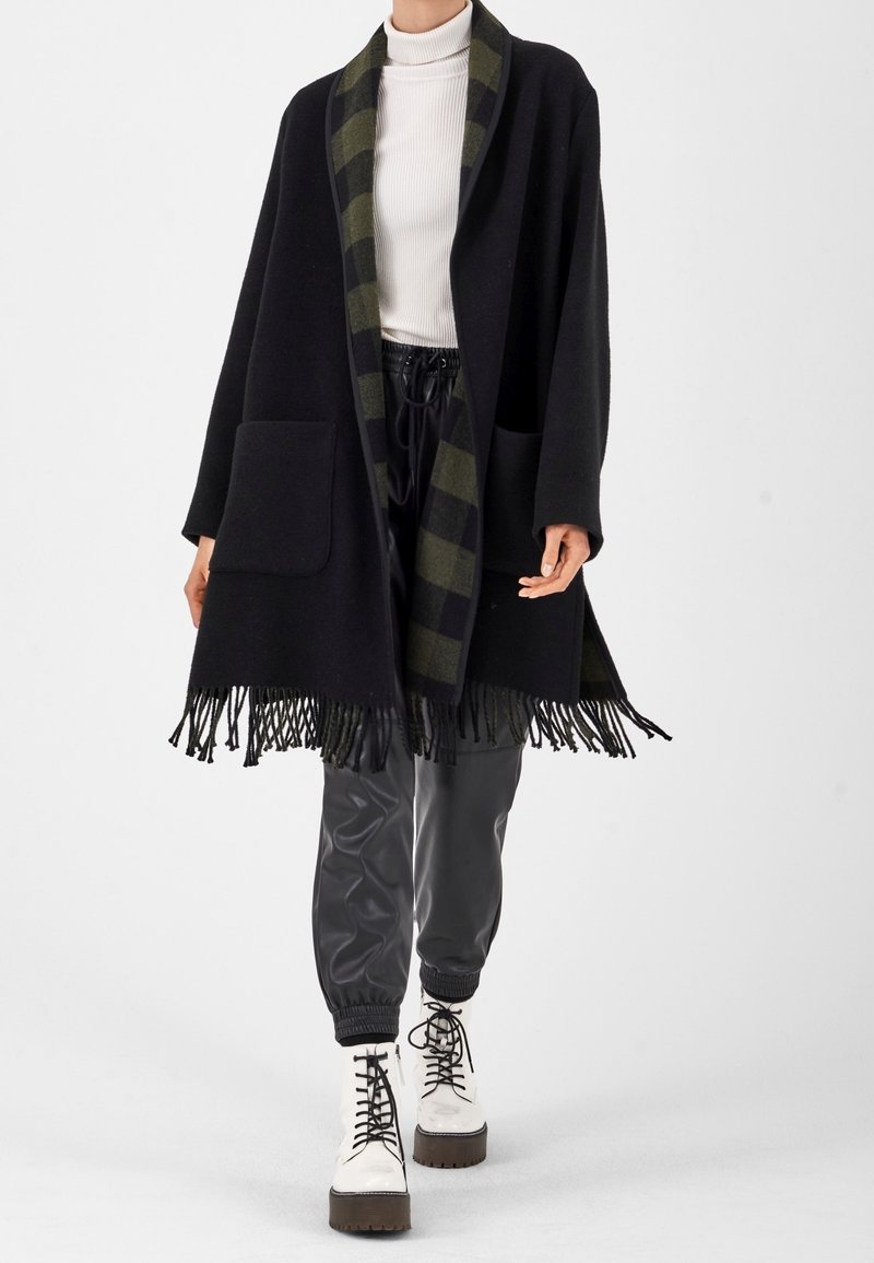 Femme portant un châle noir à franges avec doublure à carreaux verts, un col roulé blanc, un pantalon noir à cordon de serrage, et des bottes compensées blanches à lacets.