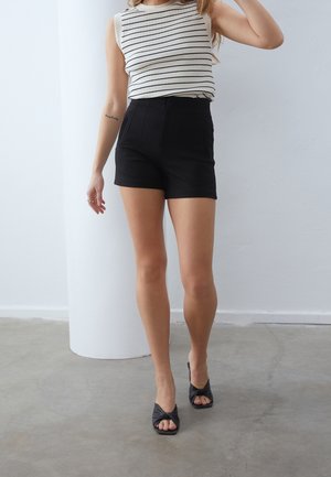 Mujer con una blusa blanca sin mangas con rayas negras, shorts negros de cintura alta y sandalias negras de tacón abierto, de pie en el interior.