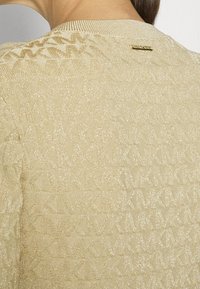 Maglione lavorato a maglia di un beige chiaro con un motivo testurizzato e rifiniture in filo metallico. Presenta un collo a coste e un piccolo etichetta con logo dorato.