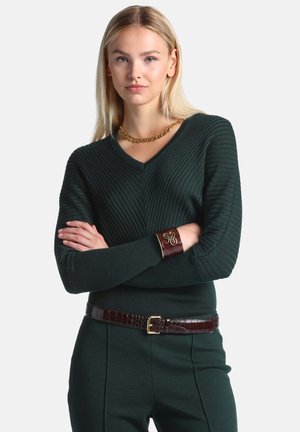 Joven mujer con cabello rubio lleva un suéter acanalado verde oscuro, pantalones a juego, un cinturón marrón, un collar de cadena dorada y una pulsera de brazalete marrón.