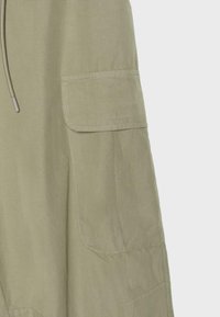 Gros plan sur une jambe de pantalon en tissu beige avec une grande poche cargo latérale à rabat et un cordon suspendu à proximité.