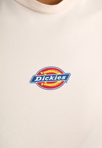 Beige tričko s kulatým výstřihem vyrobené z bavlny s hladkou texturou, s obdélníkovou červenou a žlutou logem s nápisem "Dickies" v modrém písmu.
