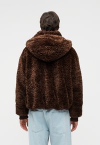 Καφέ, αφράτο σακάκι με κουκούλα και υφή εξωτερικής επιφάνειας, με ελαστικά μανίκια, φορεμένο πάνω από ανοιχτό γαλάζιο τζιν παντελόνι. Διαθέτει άνετη γραμμή και ορατές ραφές.