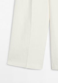 Gros plan sur la partie inférieure d'un pantalon large de couleur blanc cassé avec un pli visible sur chaque jambe, sur fond blanc.