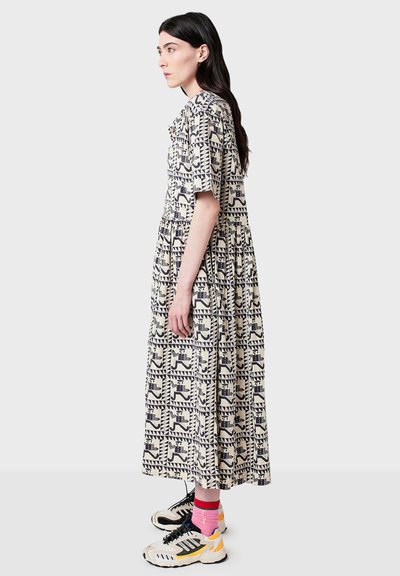 Jurk met een geometrisch patroon in beige en marineblauw; korte mouwen, geplooide taille en een zoom tot op de enkels. Gecombineerd met sportieve sneakers en sokken.