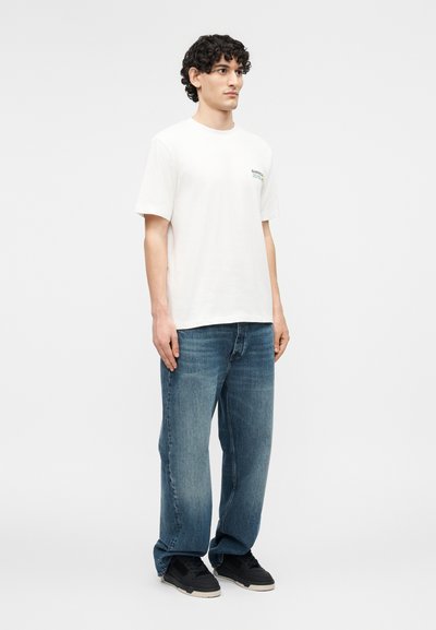 Νέος άνδρας με κατσαρά μαλλιά που φοράει λευκό κοντομάνικο μπλουζάκι, φαρδιά μπλε τζιν και μαύρα αθλητικά παπούτσια, στέκεται μπροστά σε απλό φόντο.