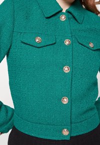 Türkise cropped Jacke aus strukturiertem Stoff, mit einem kragenförmigen Ausschnitt, zwei Brusttaschen und dekorativen goldfarbenen Knöpfen.