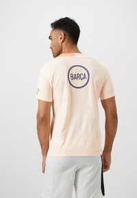 Man wearing light peach sports shirt with "BARÇA" logo on the back and white shorts, standing against a plain white background.