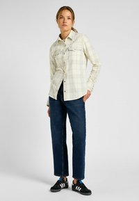 Vaaleanbeige ruudullinen paita, jossa on kaksi rintataskua, napitus ja pitkät hihat, yhdistetty tummansinisiin suoriin farkkuihin ja mustiin lenkkareihin.