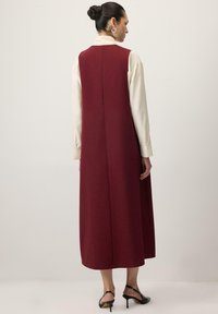 Omarantna brezrokavska obleka z dolgim, ravnim krojem, zapletenim šivom na hrbtu, nošena preko svetle bluze; obuta s črnimi sandali s peto.