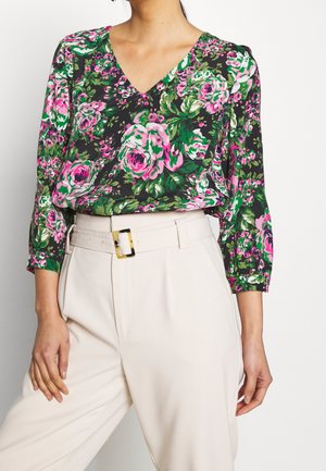 Kobieta w zielonej, kwiatowej bluzce z długim rękawem, ozdobionej różowymi kwiatami, wpuszczonej w kremowe, wysokogabarytowe spodnie z paskiem.