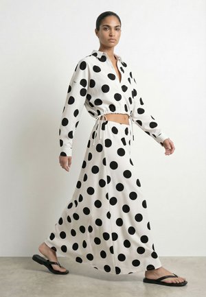 Woman walking in white cropped blouse and long skirt with large black polka dots, wearing black flip-flops, against plain background.