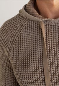 Nahaufnahme einer Person, die einen braunen, strukturierten Strickhoodie mit Kordeln trägt, wobei die Schulter, der Kragen und der obere Brustbereich sichtbar sind.