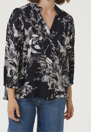 Femme portant un chemisier noir avec un motif floral gris et blanc et un jean denim bleu, debout sur un fond clair uni.